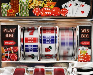 A close-up view of a slot machine with vibrant colors and various gambling symbols such as dice, coins, and playing cards. The reels display a combination of symbols, including bars, cherries, and the number seven. The chrome exterior and colorful details convey the theme of casino and gambling.