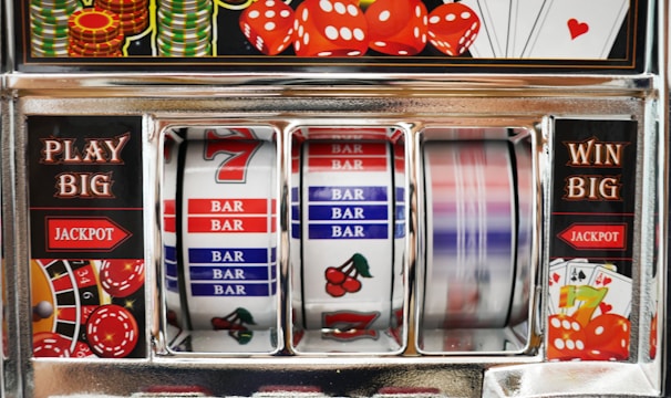 A close-up view of a slot machine with vibrant colors and various gambling symbols such as dice, coins, and playing cards. The reels display a combination of symbols, including bars, cherries, and the number seven. The chrome exterior and colorful details convey the theme of casino and gambling.