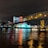 A brightly lit casino with neon signs and red lights is reflected in a river. A bridge stretches across the water, with an urban skyline visible in the background against an overcast night sky.