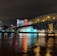 A brightly lit casino with neon signs and red lights is reflected in a river. A bridge stretches across the water, with an urban skyline visible in the background against an overcast night sky.