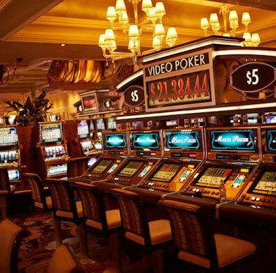 A casino floor featuring rows of brightly lit video poker machines. The area is illuminated by elegant chandeliers, with a display board showing a large sum of money, suggesting a possible jackpot. The rich decor includes luxurious carpets and comfortable seating, adding to the opulent atmosphere.
