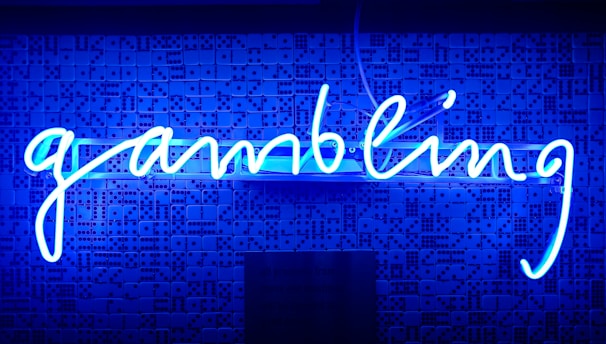 A bright blue neon sign spells out the word 'gambling' in cursive script. The background features a textured wall covered in domino tiles, also illuminated by the blue light, creating a vivid and visually striking scene.