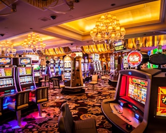 A vibrant casino interior filled with rows of colorful slot machines, each illuminated with bright lights and displays. The ceiling is adorned with elegant chandeliers and golden drapes, creating a luxurious atmosphere. The patterned carpet adds to the elaborate design of the space.