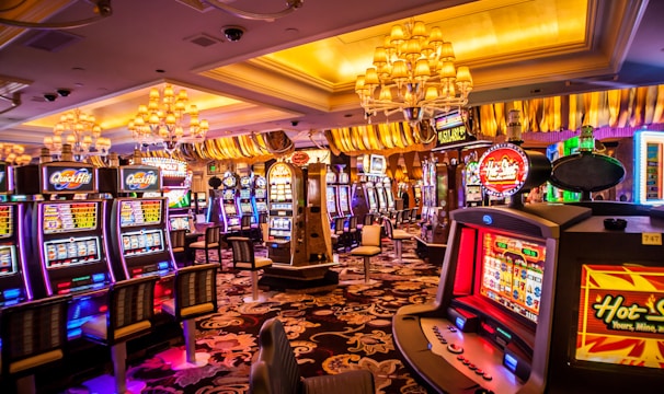 A vibrant casino interior filled with rows of colorful slot machines, each illuminated with bright lights and displays. The ceiling is adorned with elegant chandeliers and golden drapes, creating a luxurious atmosphere. The patterned carpet adds to the elaborate design of the space.