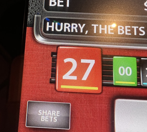 A digital display screen showing a casino betting interface, with text prompting to hurry bets and a highlighted number 27 in red. There are options for betting amounts and a 'share bets' button.